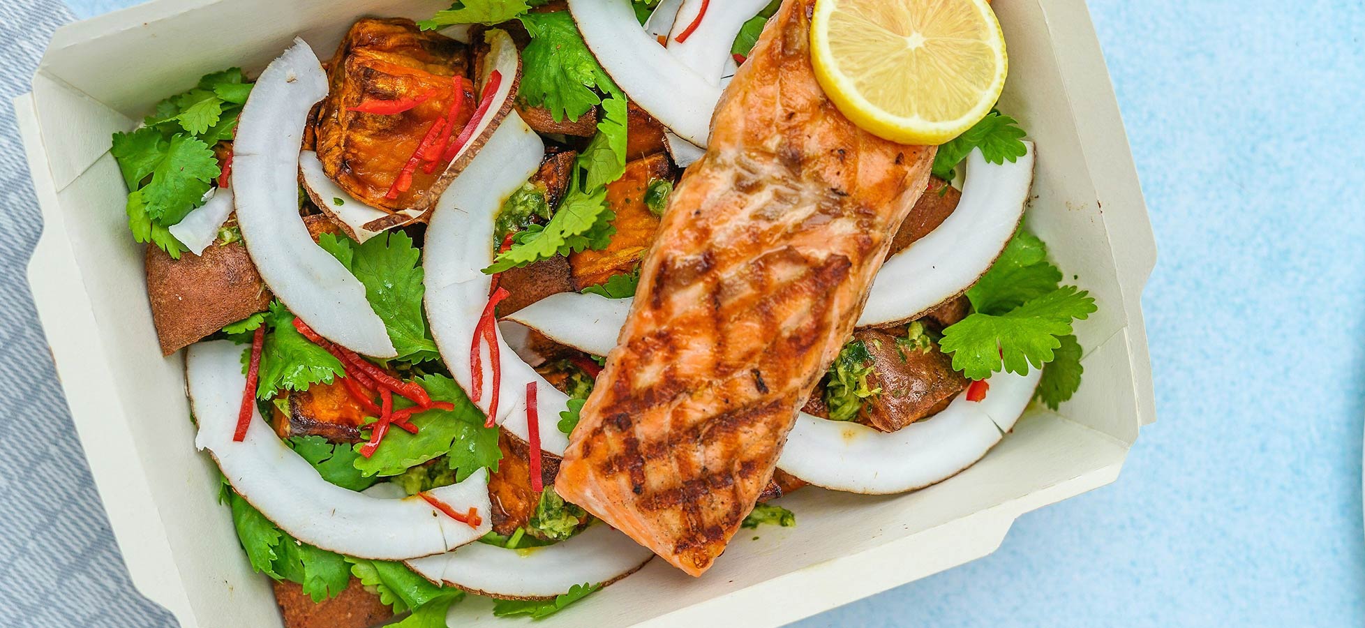 Ein Gericht mit gegrilltem Lachsfilet, garniert mit einer Zitronenscheibe. Um den Lachs liegen frische Kräuter, Kokosnussstreifen und rote Chilis auf einer Basis aus braunem Brot. Die Zutaten sind ansprechend angeordnet in einem weißen Behälter.