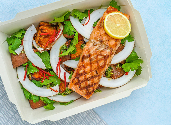 Ein Gericht in einer Pappschale mit gegrilltem Lachsfilet, garniert mit einer Zitronenscheibe. Umgeben von frischen Kräutern, darunter Koriander, sowie Kokosnuss- und Paprikastücken. Die farbenfrohe Anordnung betont die Frische der Zutaten.