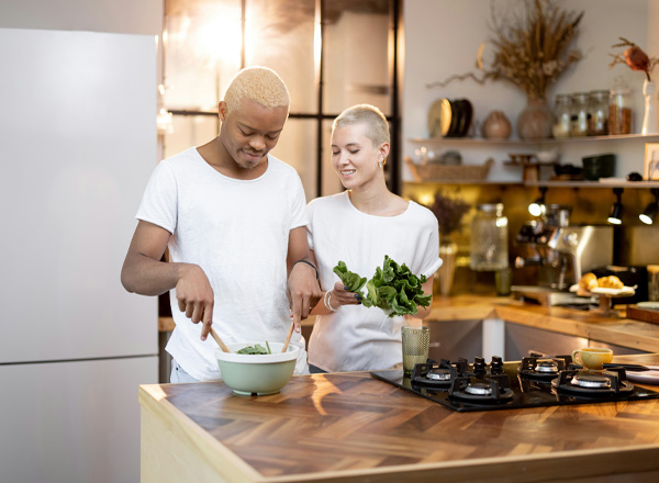 Zwei Personen bereiten in einer modernen Küche eine Mahlzeit vor. Eine Person rührt in einer Schüssel mit Salat, während die andere frisch geschnittenes Gemüse hält. Beide wirken freundlich und überrascht, und es ist eine entspannte, kreative Atmosphäre zu spüren.