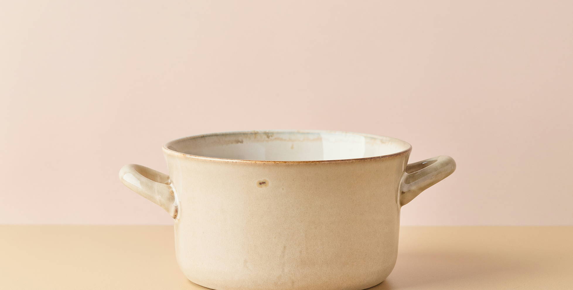 Ein beige Kochtopf mit zwei Griffen steht auf einem hellen Tisch. Der Topf hat eine schlichte, rustikale Oberfläche und eine helle Innenseite. Der Hintergrund ist in einem sanften Rosa gehalten, was eine ruhige Atmosphäre schafft.
