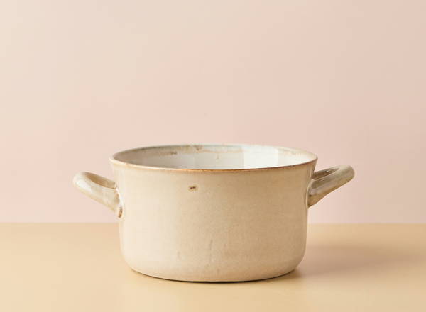Ein runder, hellbeiger Kochtopf mit zwei seitlichen Griffen steht auf einem neutralen Untergrund. Der Topf hat eine einfache Form und ist innen blau-weiß glasiert. Im Hintergrund ist eine sanfte, rosafarbene Wand zu erkennen.