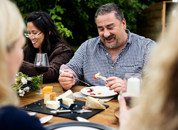 Eine Gruppe von Personen genießt ein Essen im Freien. Ein Mann in kariertem Hemd lächelt, während er Käse von einem Teller mit einer Gabel isst. Im Hintergrund ist eine Frau mit Brille zu sehen, die ein Glas Rotwein hält. Der Tisch ist mit verschiedenen Käsesorten gedeckt.