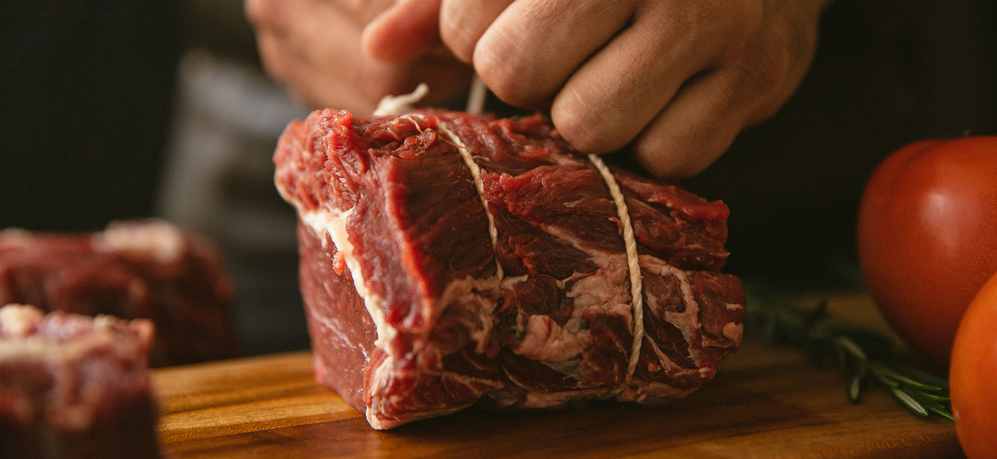 Ein Koch hält ein großes Stück rohes Fleisch auf einem Holzbrett, während er es mit Band zusammenbindet. Im Hintergrund sind weitere Fleischstücke und frische Zutaten wie Tomaten und Kräuter sichtbar.