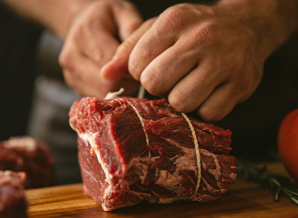 Eine Person bindet ein Stück rohes Fleisch mit Küchengarn auf einem Holzbrett. Die Hände sind aktiv, während das Fleisch straff gebunden wird, um es für die Zubereitung vorzubereiten.