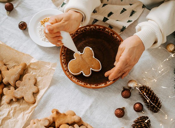 Eine Person dekoriert einen Lebkuchen mit Zuckerguss. Um sie herum liegen weitere Lebkuchen und Dekorationsmaterialien. Es gibt eine Schüssel mit bunten Streuseln auf einem Tisch mit weihnachtlicher Dekoration.
