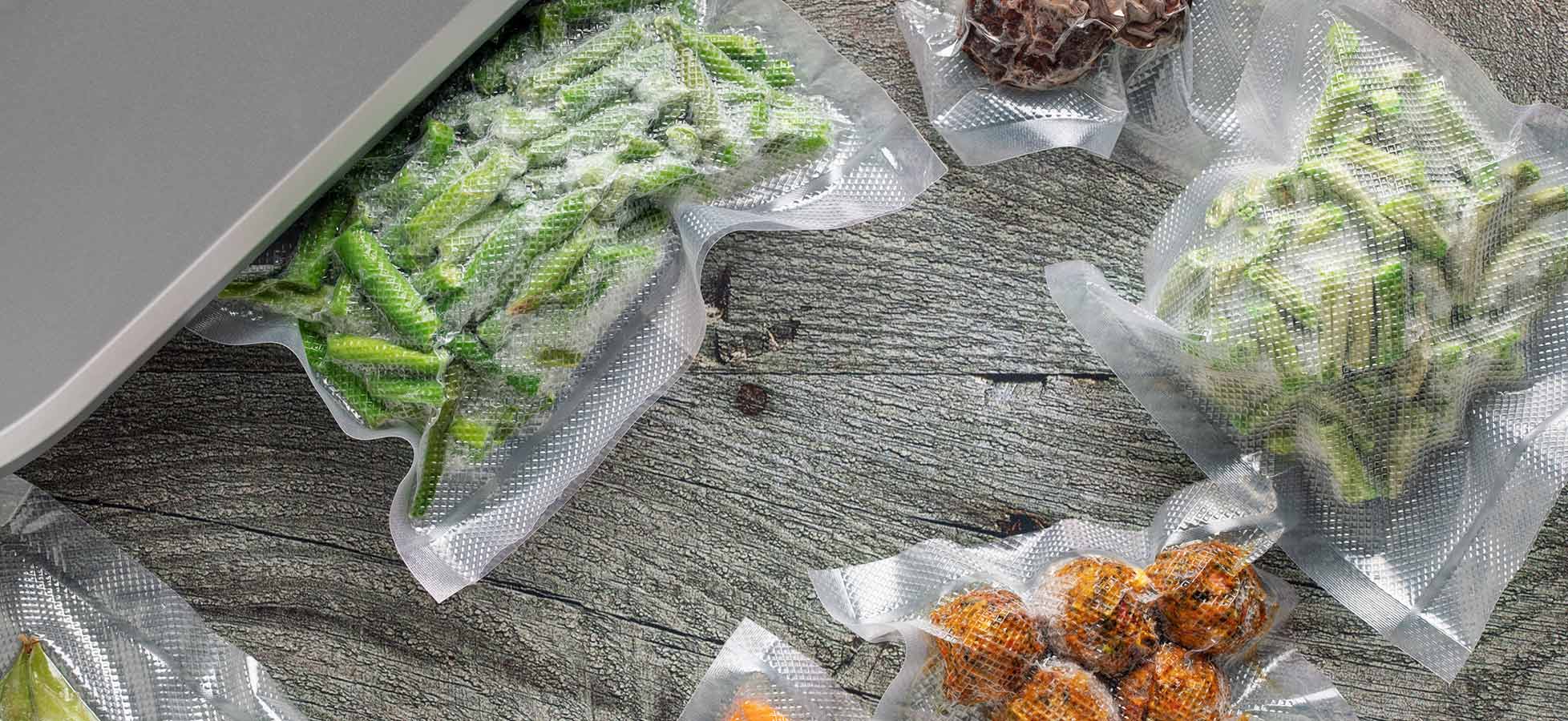 Eine Vielzahl von vakuumverpacktem Gemüse und Obst liegt auf einer Holzoberfläche. Zu sehen sind grüne Bohnen, orangefarbene Stücke und kleine braune Kugeln, die wahrscheinlich geröstete Snacks sind. Die Verpackungen sind durchsichtig, sodass der Inhalt sichtbar ist.
