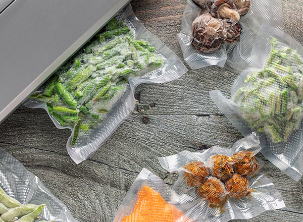 Auf einem Holztisch sind mehrere vakuumverpackte Tüten mit verschiedenen Gemüse- und Pilzsorten angeordnet. Zu sehen sind grüne Bohnen, Karotten, Zwiebeln und andere eintypische Lebensmittel. Im Hintergrund steht ein Vakuumiergerät.