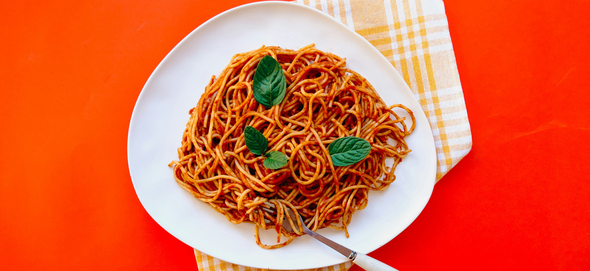 Ein Teller mit Spaghetti in Tomatensoße, garniert mit frischen Minzblättern. Der Teller ist auf einem orangefarbenen Hintergrund platziert. Ein Essbesteck ist neben dem Teller zu sehen.