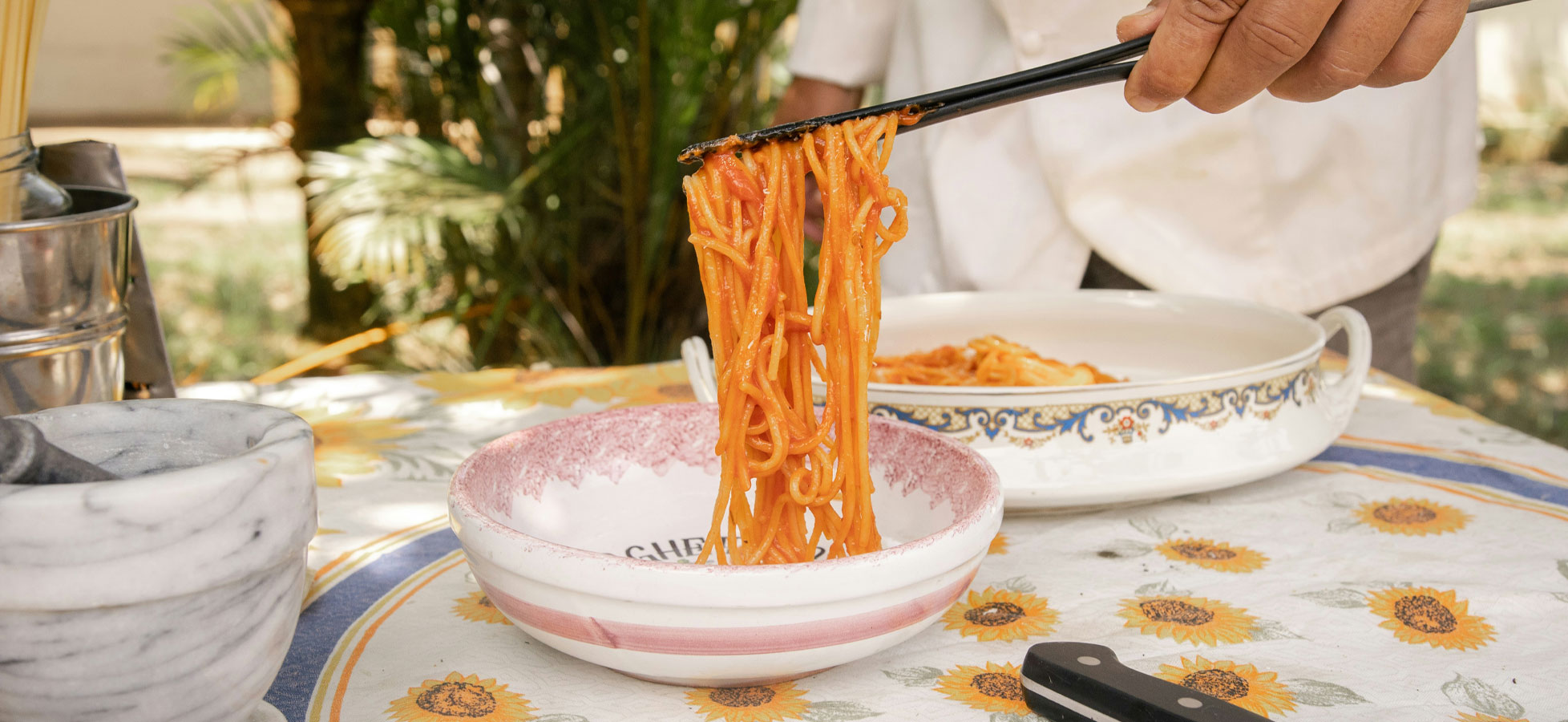 Ein Hand mit Essstäbchen hebt einen Teller Spaghetti aus einer Schüssel. Eine weiße Tischdecke mit Sonnenblumenmuster ist im Hintergrund sichtbar. Ein weiterer Teller mit Spaghetti steht neben der Schüssel. Die Szene vermittelt eine einladende Essensatmosphäre im Freien.