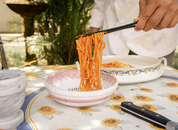 Ein Hand mit Essstäbchen hebt einen Teller Spaghetti aus einer Schüssel. Eine weiße Tischdecke mit Sonnenblumenmuster ist im Hintergrund sichtbar. Ein weiterer Teller mit Spaghetti steht neben der Schüssel. Die Szene vermittelt eine einladende Essensatmosphäre im Freien.