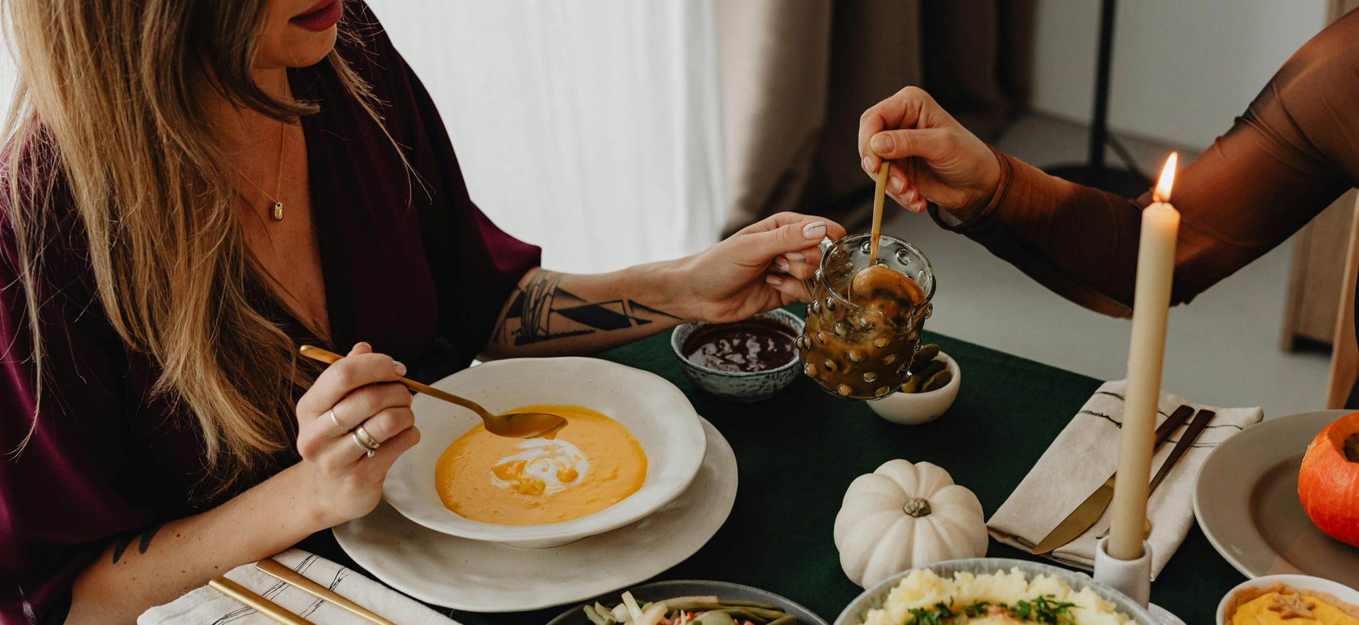 Zwei Frauen sitzen an einem Tisch und genießen ein festliches Essen. Eine Frau reicht der anderen ein Glas mit eingelegtem Gemüse. Auf dem Tisch stehen verschiedene Gerichte, darunter eine Kürbissuppe, grüne Bohnen, Kartoffelpüree und eine Schale mit Salat. Eine Kerze sorgt für gemütliche Stimmung.