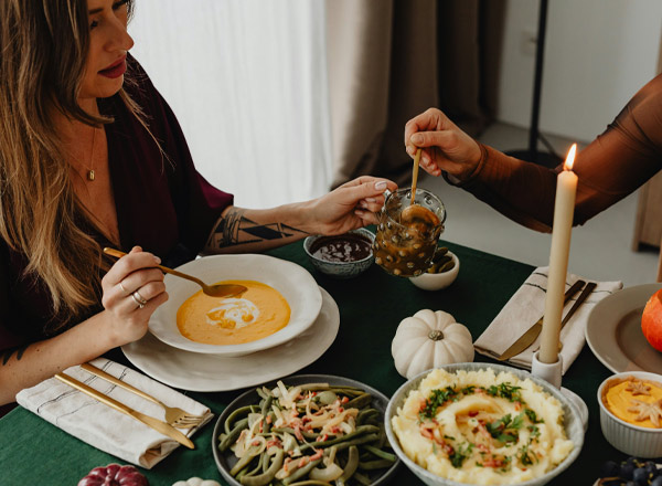 Zwei Frauen sitzen an einem Tisch und genießen ein festliches Essen. Eine Frau reicht der anderen ein Glas mit eingelegtem Gemüse. Auf dem Tisch stehen verschiedene Gerichte, darunter eine Kürbissuppe, grüne Bohnen, Kartoffelpüree und eine Schale mit Salat. Eine Kerze sorgt für gemütliche Stimmung.