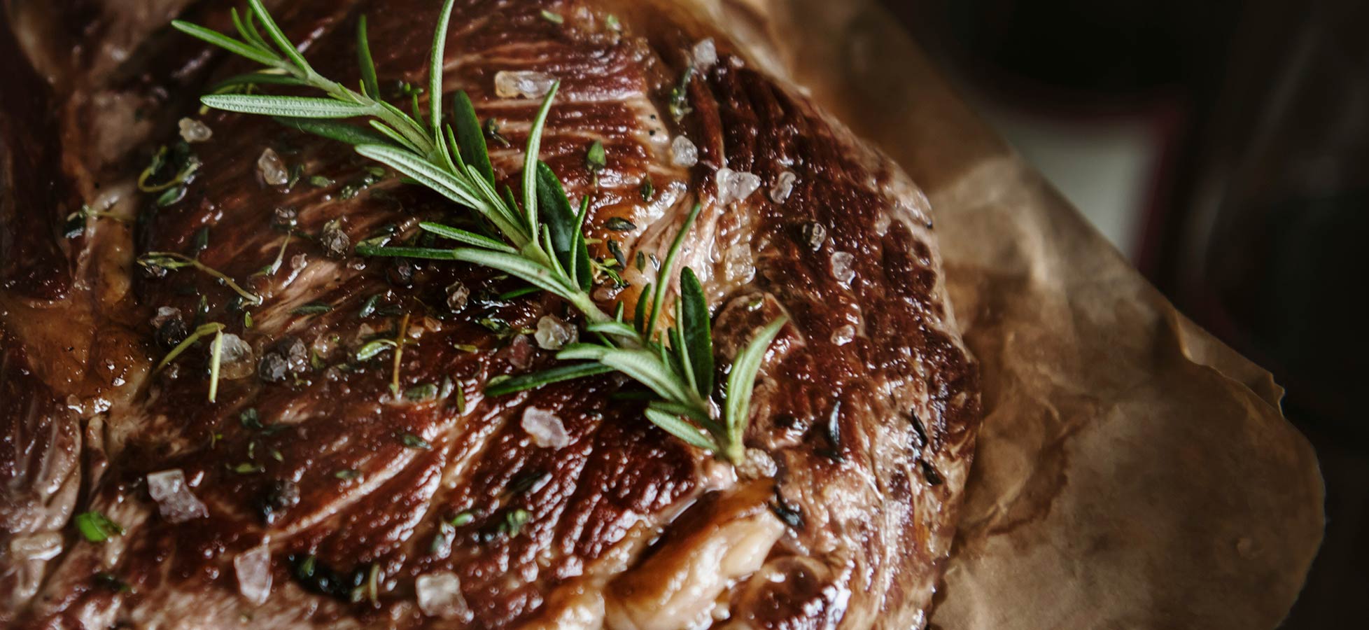 Ein saftiges Stück Fleisch liegt auf braunem Pergamentpapier. Es ist mit frischen Rosmarinzweigen und grobem Salz garniert, was den appetitanregenden Eindruck verstärkt. Die Obertfläche des Fleisches glänzt leicht und zeigt eine verführerische Bräunung.