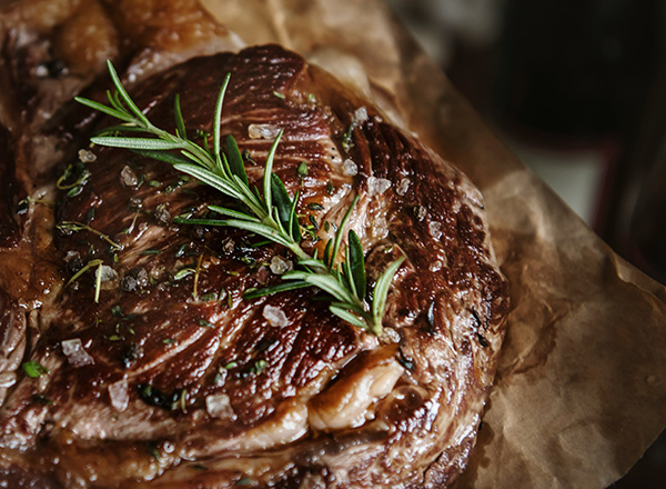 Ein saftiges, gebratenes Stück Fleisch liegt auf braunem Papier. Es ist mit grobem Salz bestreut und mit frischen Rosmarin- und Thymianzweigen garniert, was für ein herzhaftes Aroma sorgt.