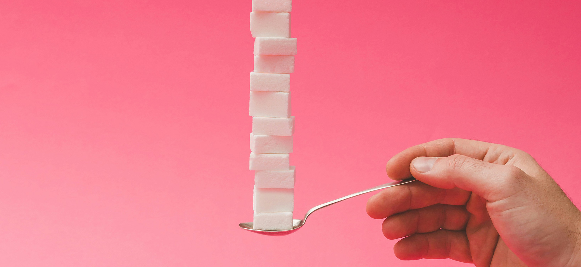Eine Hand hält einen Löffel, auf dem eine hohe Säule aus weißen Zuckerstückchen balanciert. Der Hintergrund ist pink. Die Darstellung zeigt die Anhäufung von Zucker, was auf den Zuckerkonsum hinweisen könnte.