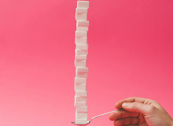 Eine Hand hält einen Löffel, auf dem eine hohe Säule aus weißen Zuckerstückchen balanciert. Der Hintergrund ist pink. Die Darstellung zeigt die Anhäufung von Zucker, was auf den Zuckerkonsum hinweisen könnte.