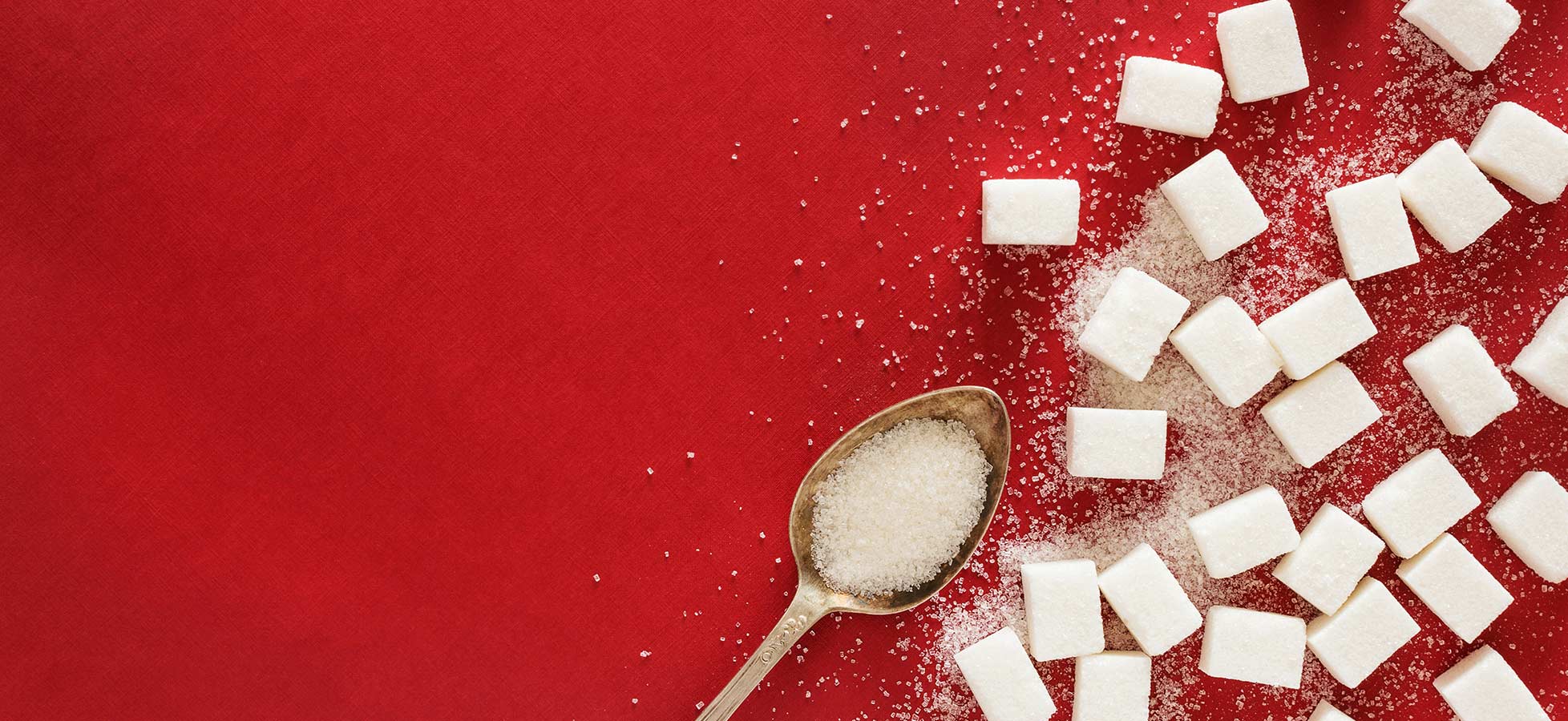 Ein roter Hintergrund zeigt verstreute Würfelzucker und eine Löffel mit Kristallzucker. Die weißen Zuckerwürfel sind unregelmäßig angeordnet, während der Löffel am unteren Rand des Bildes liegt.