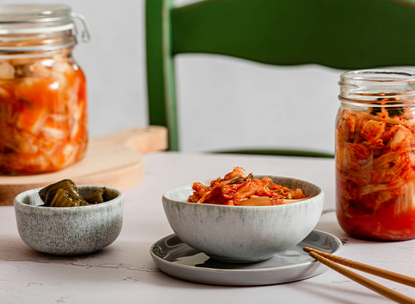 Schüssel mit Kimchi auf einem Teller, neben einer kleinen Schüssel mit einer grünen Beilage. Im Hintergrund ein Glas mit mehr Kimchi. Essstäbchen liegen auf dem Teller. Der Tisch hat eine helle Oberfläche.