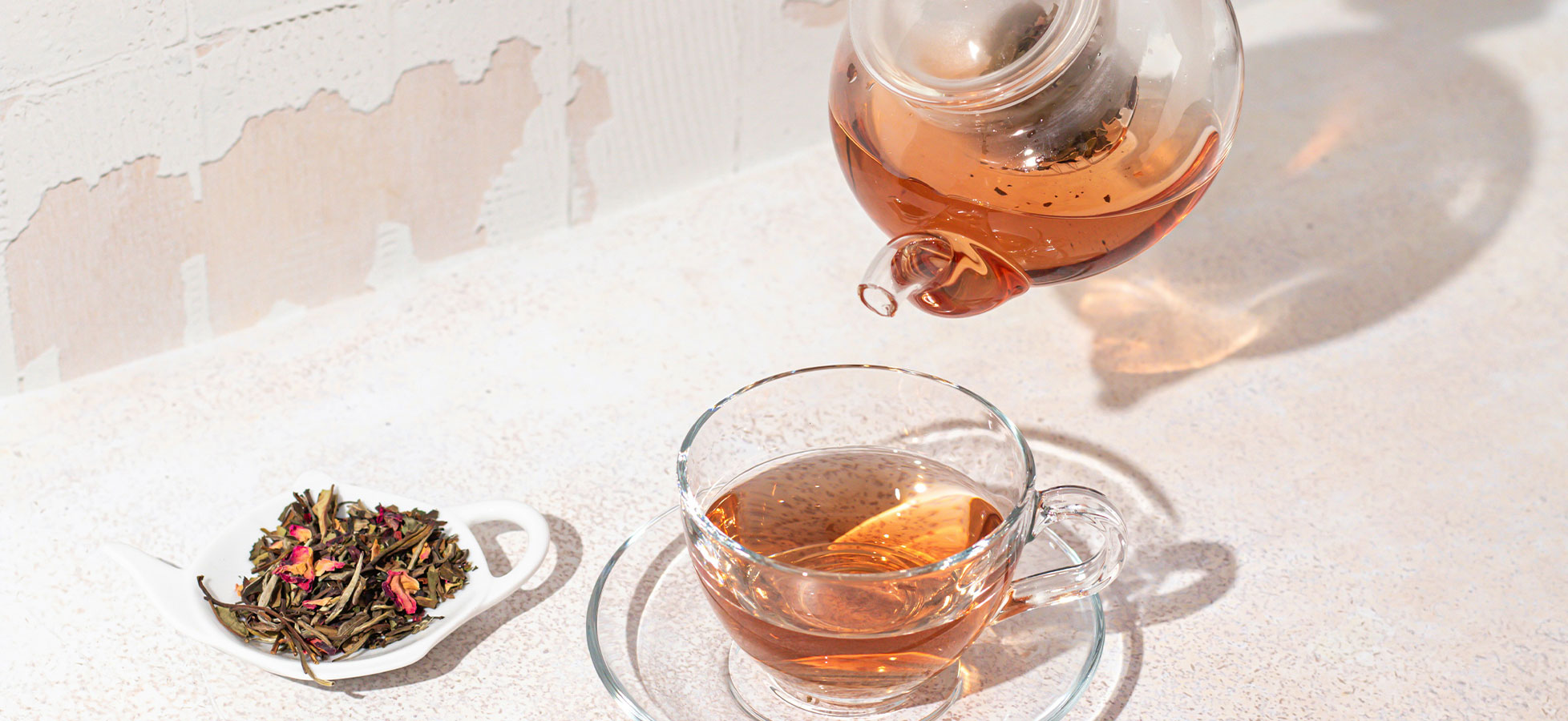 Eine Teekanne giesst frisch gebrühten Tee in eine durchsichtige Tasse, die auf einem Unterteller steht. Neben der Tasse liegt eine kleine Portion loser Tee auf einem weißen Teller.