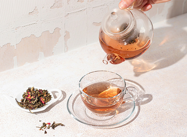 Eine Hand gießt aromatischen Tee aus einer durchsichtigen Teekanne in eine klare Tasse. Neben der Tasse liegt eine kleine Schale mit losem Tee und getrockneten Blüten.