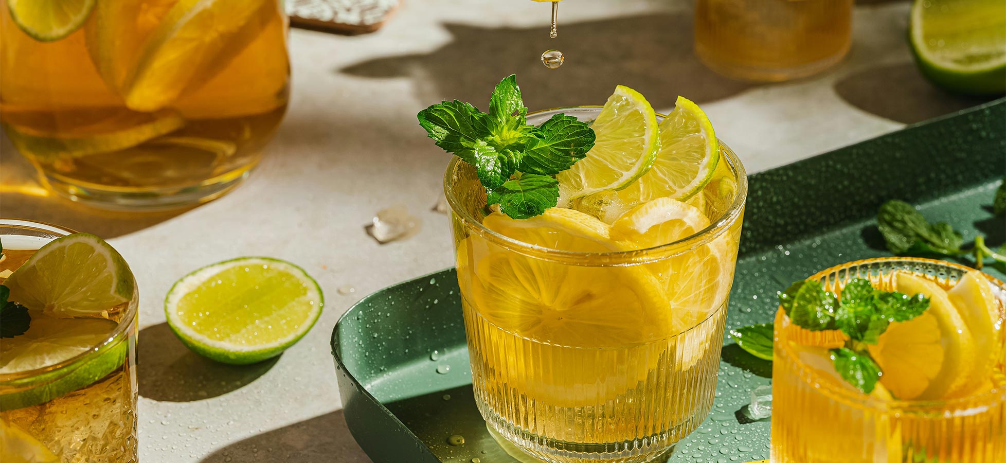 Ein Glas mit kühlem Getränk, gefüllt mit Zitronenscheiben und frischen Minzblättern, steht auf einem Tablett. Im Hintergrund sind weitere Gläser und eine Kanne mit ähnlichem Inhalt zu sehen. Limettenscheiben liegen verteilt auf der Oberfläche.