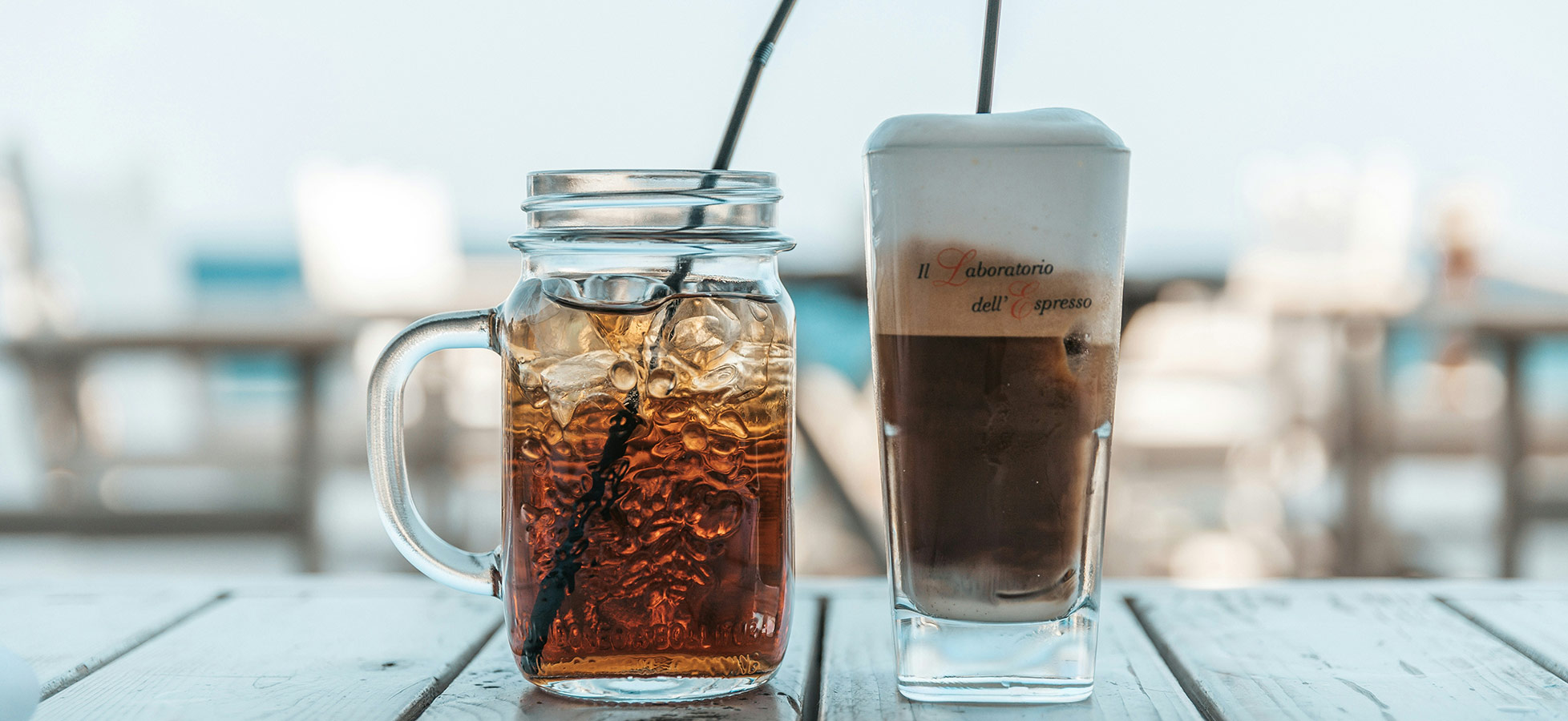 Zwei Getränke stehen auf einem Tisch: Ein großes Glas mit Eistee und Eiswürfeln, neben einem schlanken Glas mit einer schaumigen Kaffeekreation. Beide haben einen Strohhalm und vermitteln ein entspanntes Ambiente.