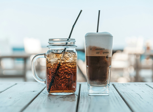 Zwei Getränke stehen auf einem Holztisch. Links ein Glas mit einem dunklen Erfrischungsgetränk und einem Strohhalm. Rechts ein hohes Glas mit Kaffee, bedeckt von einer Schicht Sahne. Im Hintergrund ist eine unscharfe Bokeh-Ansicht eines hellen, sonnigen Raumes.