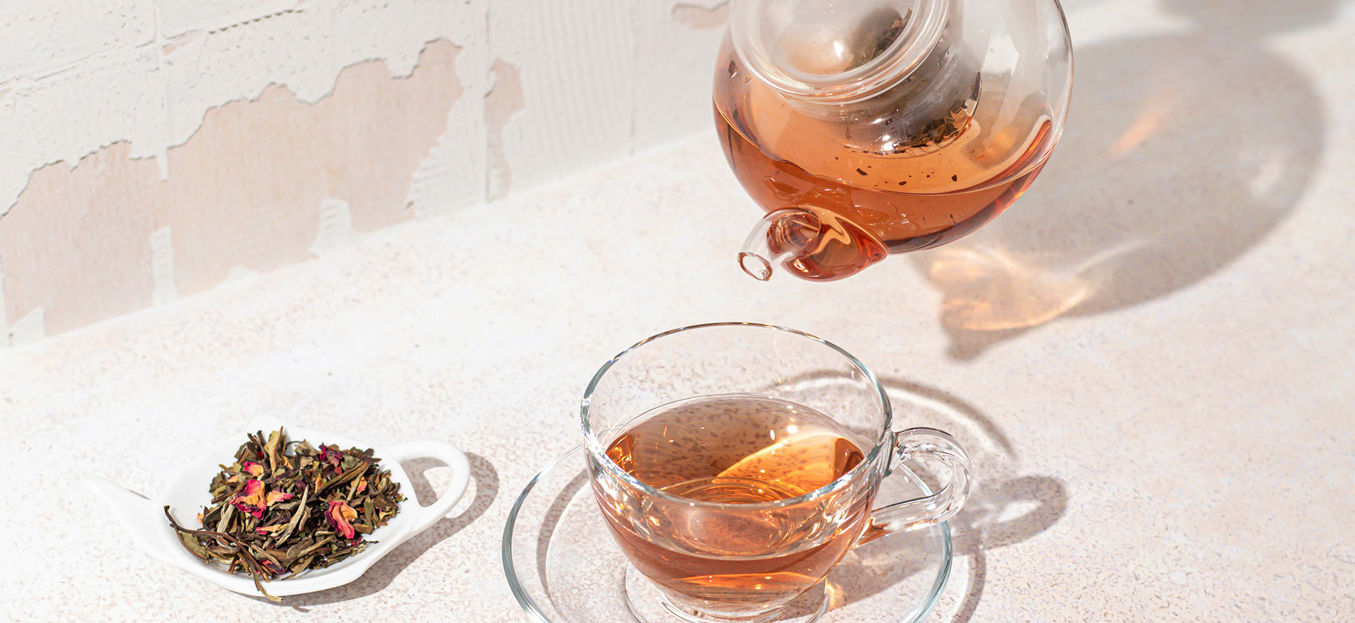 Eine Teekanne gießt warmen, klaren Tee in eine gläserne Tasse. Neben der Tasse liegt ein kleines Schälchen mit losem Tee und getrockneten Blütenblättern. Der Hintergrund zeigt eine helle, strukturierte Oberfläche.