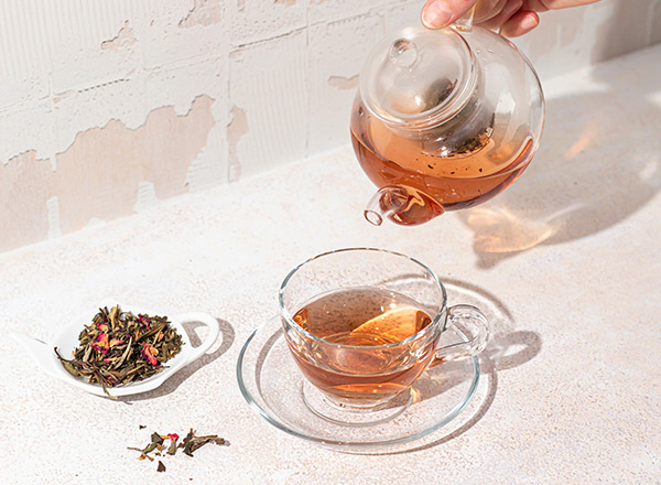 Eine Teekanne gießt aromatischen Tee in eine durchsichtige Teetasse, die auf einer Untertasse steht. Neben der Tasse befindet sich eine kleine Schale mit losem Tee, der mit Blütenblättern verziert ist. Der Hintergrund ist hell und schlicht.
