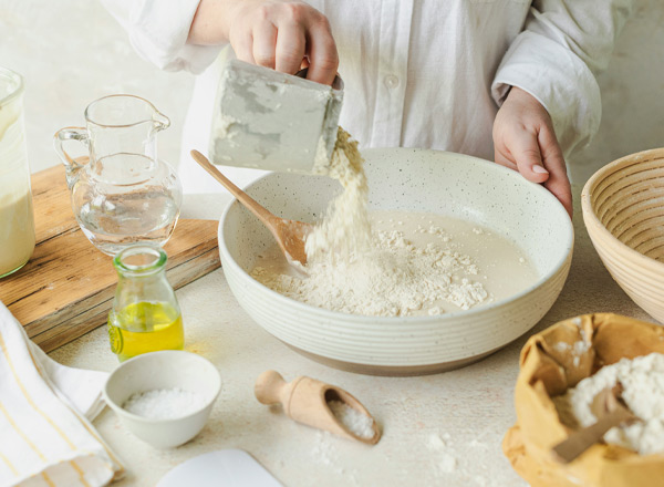 Eine Hand gießt Mehl aus einem Behälter in eine große Schüssel, die teilweise mit einer Flüssigkeit gefüllt ist. Rund um die Schüssel sind verschiedene Kochutensilien und Zutaten wie Öl, Wasser und Salz zu sehen, die für die Zubereitung benötigt werden.