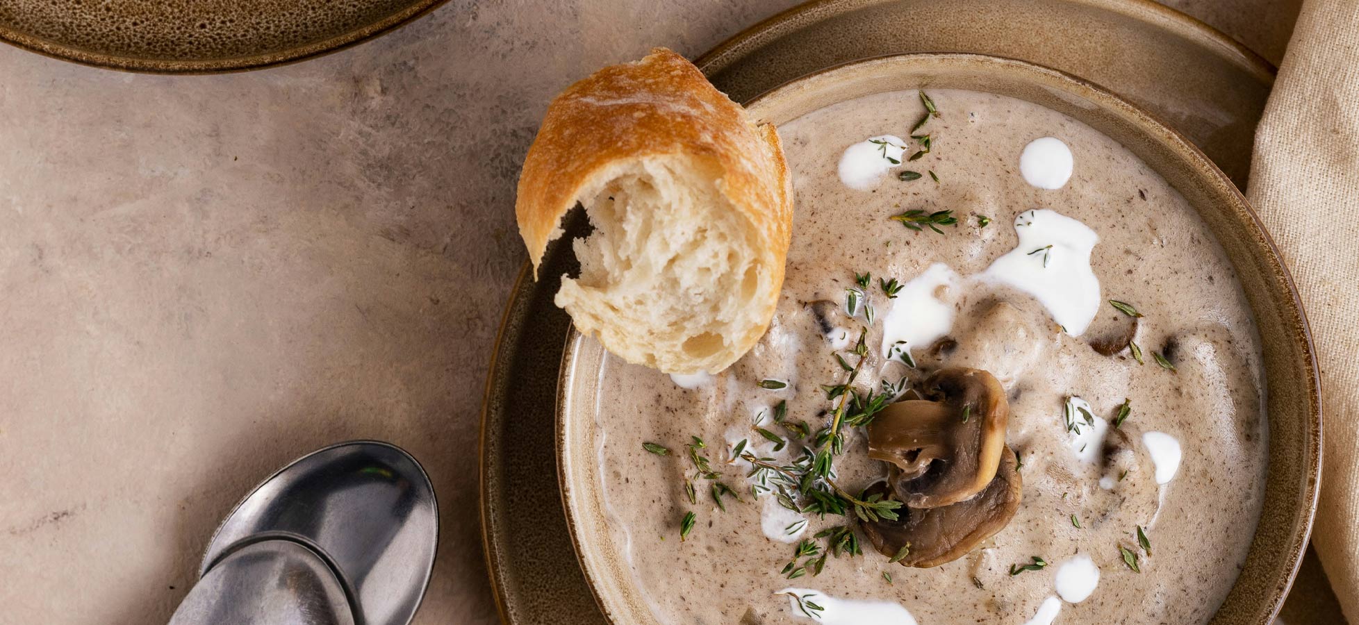Eine Schüssel mit cremiger Pilzsuppe, garniert mit frischem Thymian und einem Klecks Sahne. Neben der Schüssel liegt ein Stück knuspriges Brot. Der Hintergrund ist neutral gehalten, sodass die Suppe im Mittelpunkt steht.