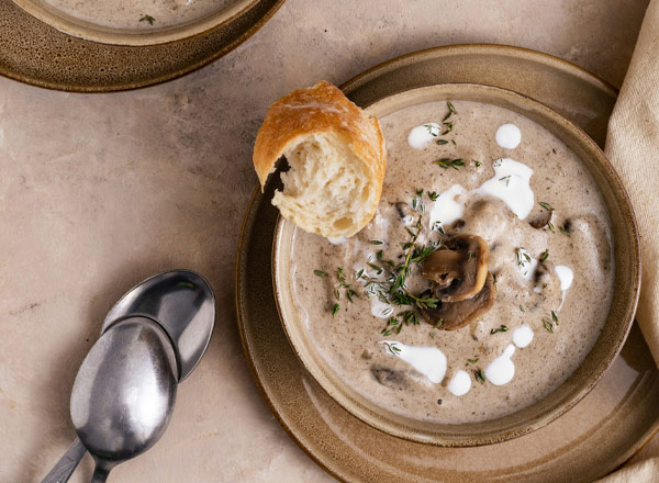 Eine Schüssel cremige Pilzsuppe, garniert mit einem Spritzer Sauerrahm und frischen Kräutern. Neben der Suppe liegt ein Stück frisches Brot. Zwei Löffel aus Metall liegen bereit, um die Suppe zu genießen.