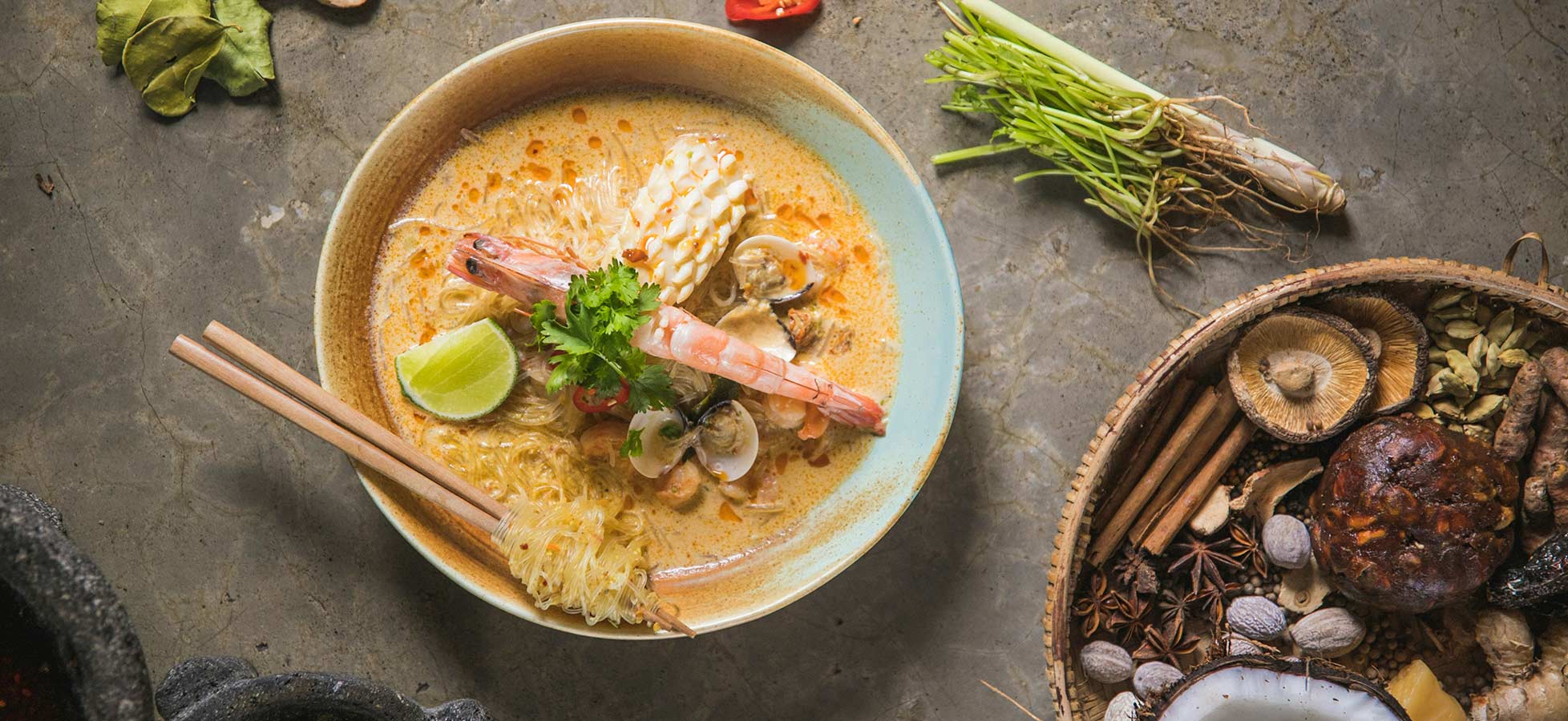 Eine Schüssel Ramen-Nudelsuppe mit Garnelen, Muscheln und frischen Kräutern. Limettenscheiben fügen Frische hinzu. Die Suppe hat eine rötliche Farbe und ist dezent mit Gewürzen angerichtet. Daneben liegen verschiedene Zutaten wie Pilze und Gewürze auf einem rustikalen Tisch.