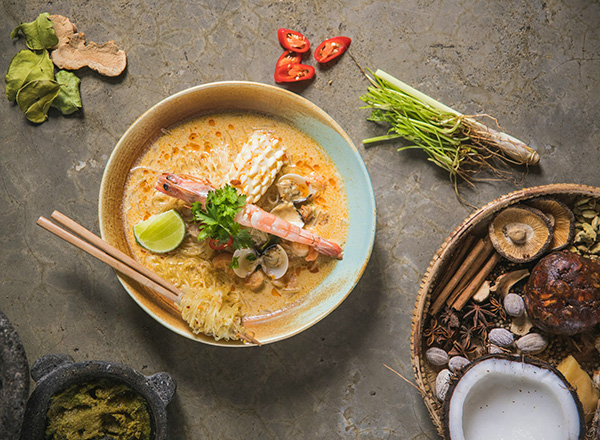 Eine Schüssel mit aromatischer Ramen-Nudelsuppe ist zu sehen, garniert mit Garnelen, Muscheln und frischen Kräutern. Limettenscheiben und Chili ergänzen das Gericht. Neben der Schüssel liegen Essstäbchen. Im Hintergrund sind verschiedene Gewürze und Zutaten angeordnet.