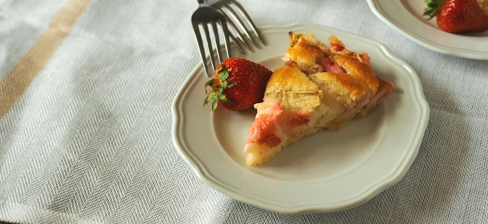 Ein Teller mit einem Stück Kuchen, das mit frischen Erdbeeren garniert ist. Eine Gabel liegt neben dem Kuchen, der eine goldene, knusprige Kruste und eine fruchtige Füllung hat. Die Hintergrundoberfläche ist hell und texturiert.