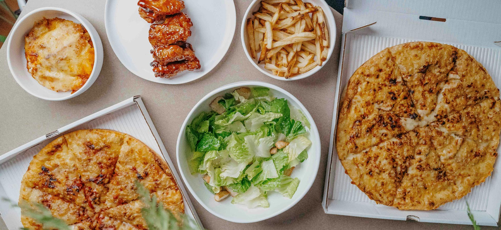 Eine Übersicht von verschiedenen Gerichten auf einem Tisch. Es gibt zwei Pizzen, eine Schüssel mit Salat, frittierte Kartoffeln und eine Portion gegrillte Hähnchenflügel in einer Schüssel. Einige Speisen befinden sich in Pizzakartons.