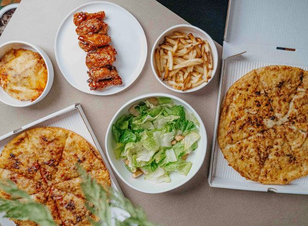 Eine Übersicht von verschiedenen Gerichten auf einem Tisch. Es gibt zwei Pizzen, eine Schüssel mit Salat, frittierte Kartoffeln und eine Portion gegrillte Hähnchenflügel in einer Schüssel. Einige Speisen befinden sich in Pizzakartons.