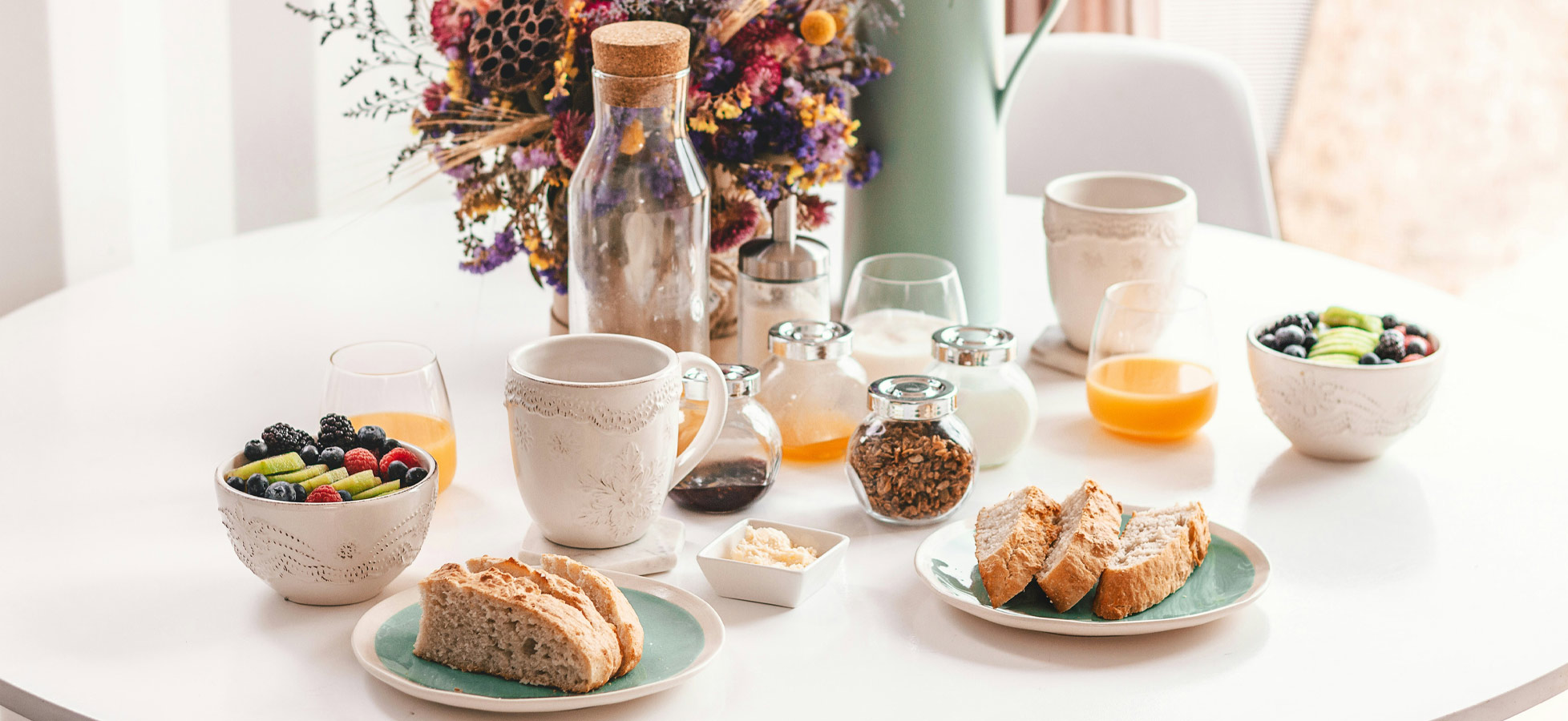 Auf einem Tisch steht ein Frühstücksarrangement: zwei Tassen, verschiedene Fruchtschalen mit Beeren und Äpfeln, eine Schüssel mit Müsli, mehrere Gläser Säfte und Brot auf einem Teller. In der Mitte befindet sich eine Glasflasche mit Korkverschluss und ein dekorativer Blumenstrauß.