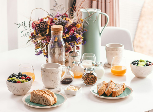 Ein Tisch ist mit einer Fülle von Frühstücksartikeln gedeckt: verschiedene Brotsorten auf Tellern, Obst in einer Schüssel, Gläser mit Getränken und kleinen Gefäßen mit Marmelade und Granola. Im Hintergrund steht eine blühende Vase.