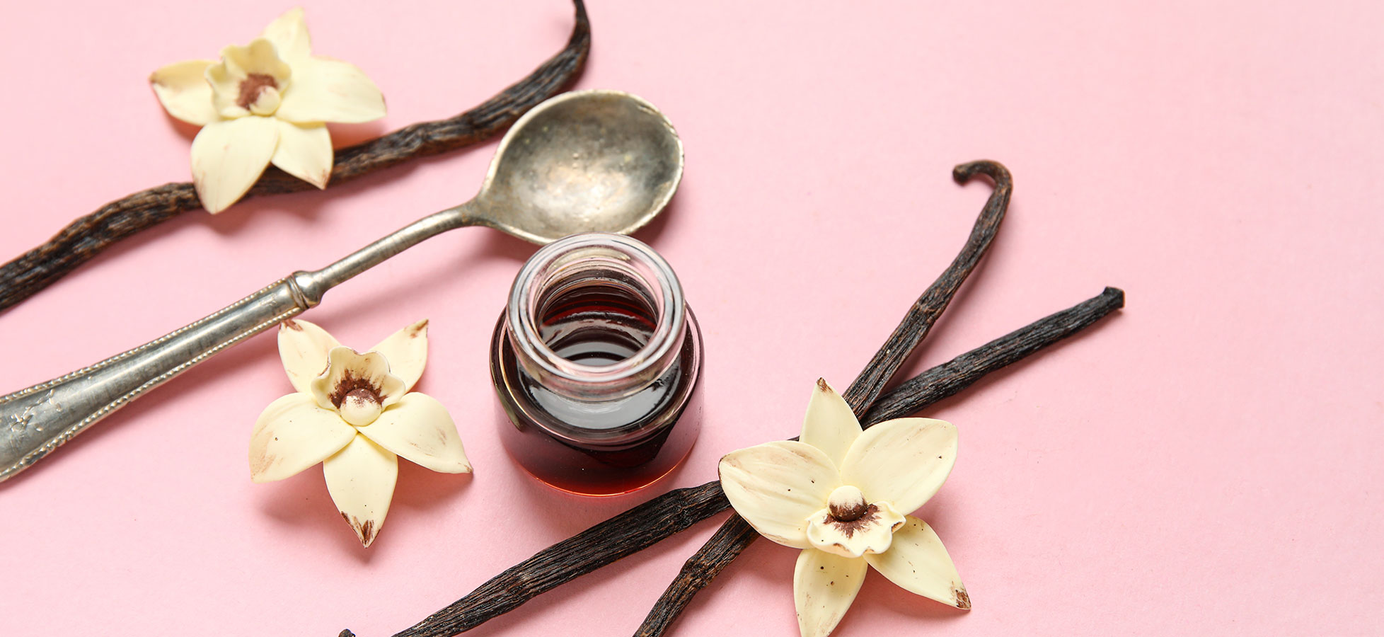 Ein Glas Fläschchen mit dunkler Flüssigkeit steht auf einem rosa Hintergrund, umgeben von Vanilleschoten und hellen Vanilleblüten. Ein Löffel aus Silber liegt ebenfalls daneben. Die Anordnung vermittelt ein Gefühl von Eleganz und Genuss.