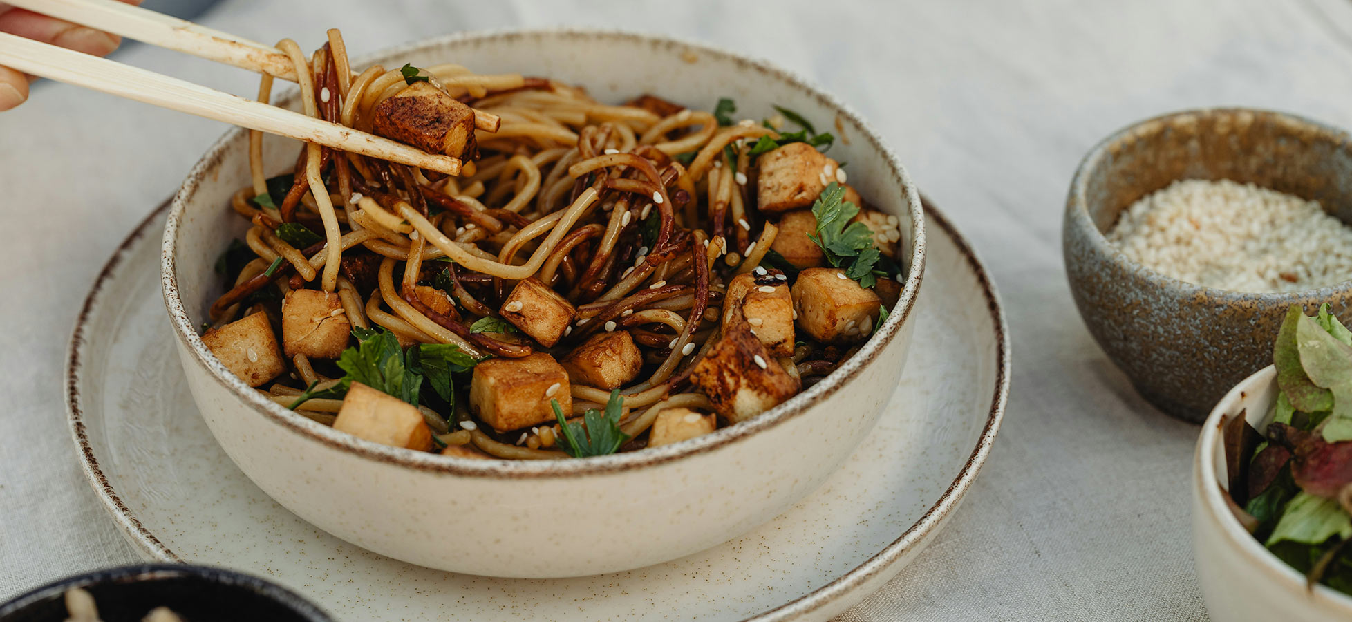 Eine Schüssel mit gebratenen Nudeln, Tofu und frischen Kräutern wird mit Essstäbchen gehalten. Die Nudeln sind mit einer dunklen Soße überzogen und mit Sesam bestreut. Die Schüssel steht auf einem weißen Teller, daneben befinden sich kleine Schalen mit Sesam und Salat.