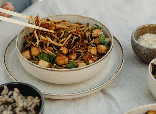 Eine Schüssel mit gebratenen Nudeln, Tofu und frischen Kräutern, während eine Hand mit Essstäbchen eine Portion herausnimmt. Im Hintergrund stehen kleinere Schalen mit weiteren Zutaten. Das Gericht liegt auf einer neutralen Tischdecke.