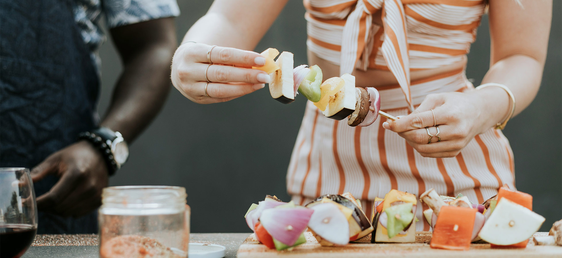Eine Hand hält einen bunten Obstspieß mit verschiedenen Früchten, während im Hintergrund ein weiterer Mensch steht. Auf einem Tisch sind mehrere Obststückchen und ein Glas zu sehen. Es vermittelt eine Atmosphäre des gemeinsamen Kochens oder Essens.