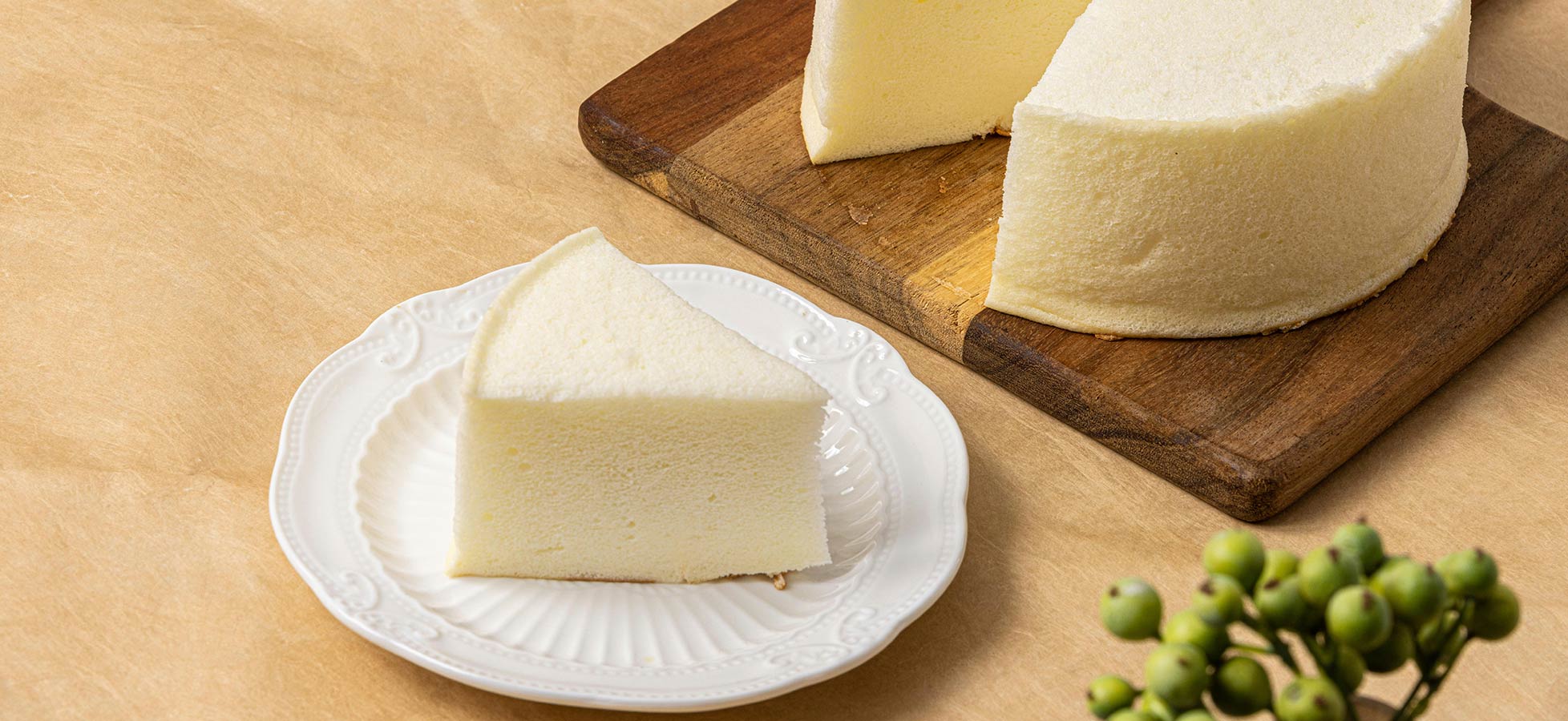 Ein Stück weicher Käse liegt auf einem weißen Teller, neben einem größeren Käseblock auf einem Holzbrett. Im Vordergrund sind grüne Trauben sichtbar. Der Hintergrund ist neutral und schlicht.