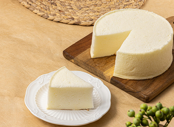 Ein runder, cremiger Kuchen steht auf einem Holzbrett. Ein Stück des Kuchens ist auf einem weißen Teller, der daneben liegt. Der Kuchen hat eine glatte, helle Oberfläche und wirkt appetitlich. Der Hintergrund ist schlicht und neutral gehalten.