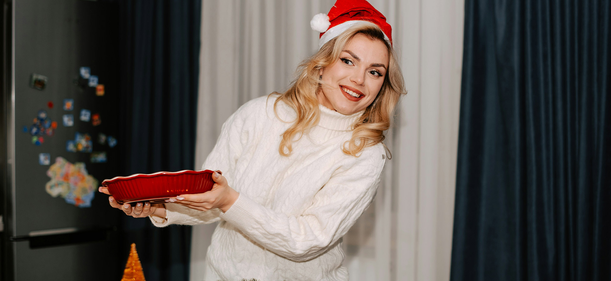 Eine lächelnde Frau trägt einen roten Teller und trägt einen knallroten Weihnachtsmann-Hut. Sie steht in einer hellen, festlichen Umgebung mit Vorhängen im Hintergrund und wirkt einladend und fröhlich.