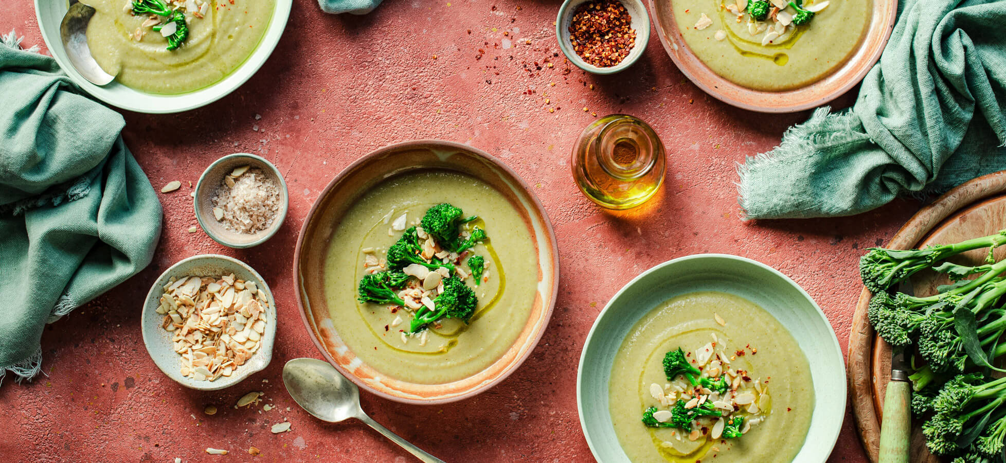Eine Auswahl an Schalen mit grüner Suppe, garniert mit Brokkoli und Mandeln, steht auf einer rustikalen Oberfläche. Neben den Schalen befinden sich kleine Schalen mit Salz, roten Pfefferflocken und Haferflocken sowie ein Fläschchen Öl und ein grünes Tuch.