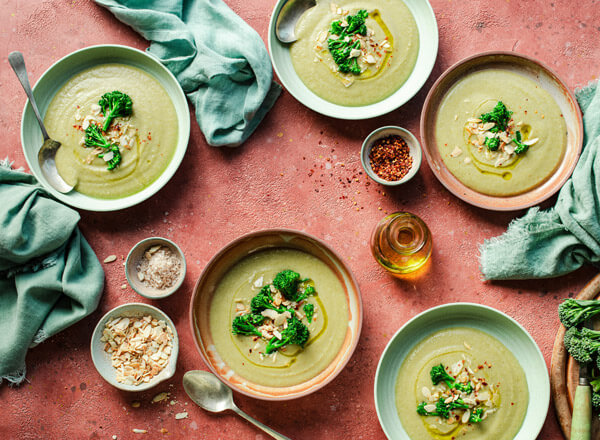 Fünf Schalen mit cremiger Brokkolisuppe stehen auf einem rustikalen Tisch. Die Suppe ist mit Brokkoliröschen und gerösteten Mandeln garniert. Um die Schalen herum sind kleine Schalen mit Gewürzen und Öl verteilt. Ein frisches, einladendes Gericht.