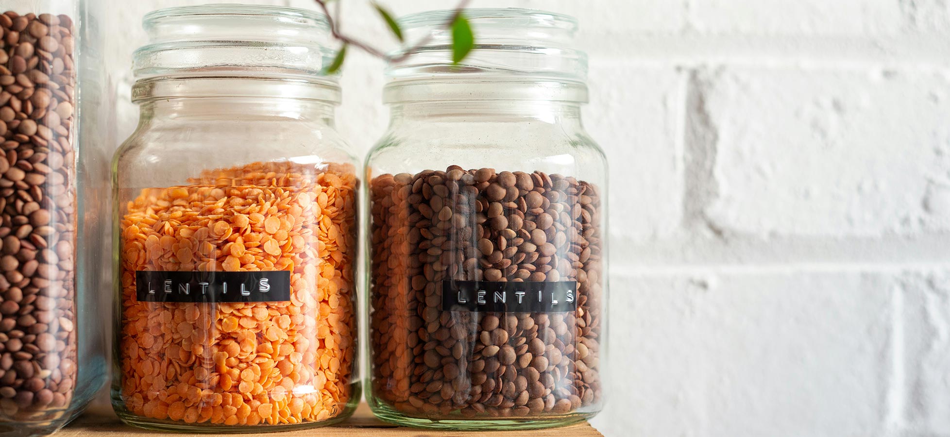 Eingelegte Gläser mit Linsen sind auf einem Holzregal angeordnet. Das Glas in der Mitte enthält orangene Linsen, während das rechte Glas braune Linsen enthält. Beide haben ein Etikett mit der Aufschrift „LENTILS“. Der Hintergrund ist eine weiße Ziegelwand.