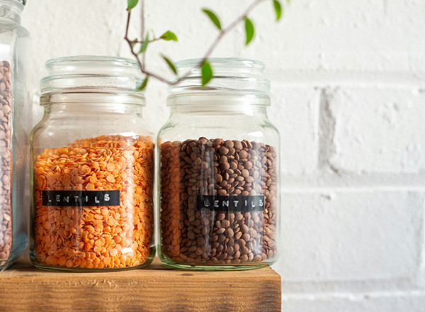 Zwei durchsichtige Glasbehälter auf einem Holzregal, einer mit orangenen Linsen und der andere mit braunen Linsen, beide beschriftet. Im Hintergrund ist eine weiße Mauer zu sehen. Ein paar grüne Blätter sind im Bild.